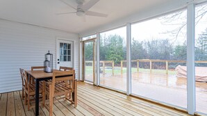 Eine überdachte Terrasse mit Holztisch und -stühlen, großen Fenstern und einem Deckenventilator.