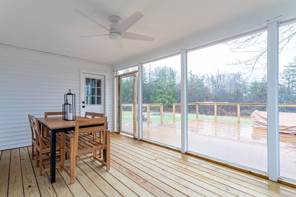 A covered deck with a wooden table and chairs, large windows, and a ceiling fan.