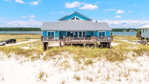 Ein Strandhaus mit blauem Dach und Holzterrasse, mit Blick auf einen Sandstrand und ein Gewässer.
