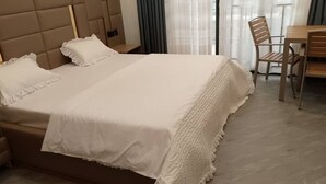 Una cama bien hecha con edredón blanco y almohadas, cabecera de madera y una mesa de madera con sillas en una habitación con un ventanal y cortinas.
