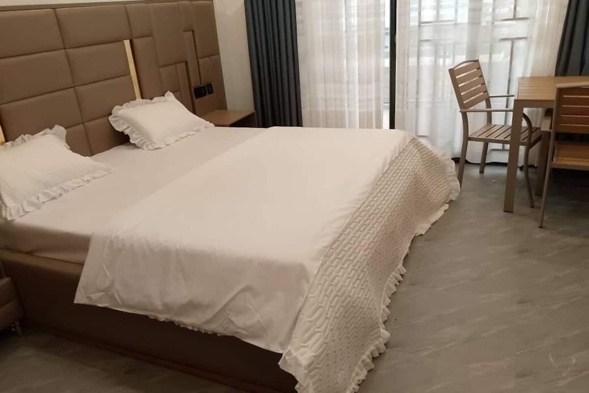 A neatly made bed with a white duvet and pillows, a wooden headboard, and a wooden table with chairs in a room with a large window and curtains.