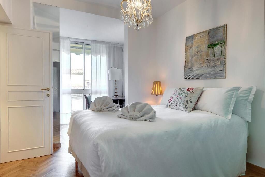 A hotel room with a large bed, a chandelier, a painting on the wall, and a door leading to another room.