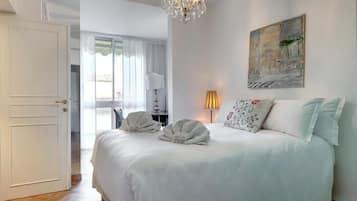 A hotel room with a large bed, a chandelier, a painting on the wall, and a door leading to another room.