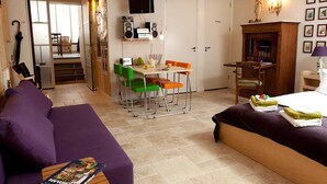 Un espace de vie compact comprenant un canapé violet, une table à manger avec des chaises, un téléviseur fixé au-dessus de la table et une cheminée ornée de tableaux encadrés sur le mur.