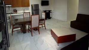 A living room with a sofa, a dining table with chairs, a kitchen area with cabinets and appliances, and a TV mounted on the wall. - Residencia Gran Coyula Orange 512B (Tonala)