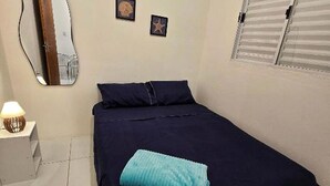 A bedroom with a bed, a nightstand with a lamp, a wall mirror, and a window with blinds.