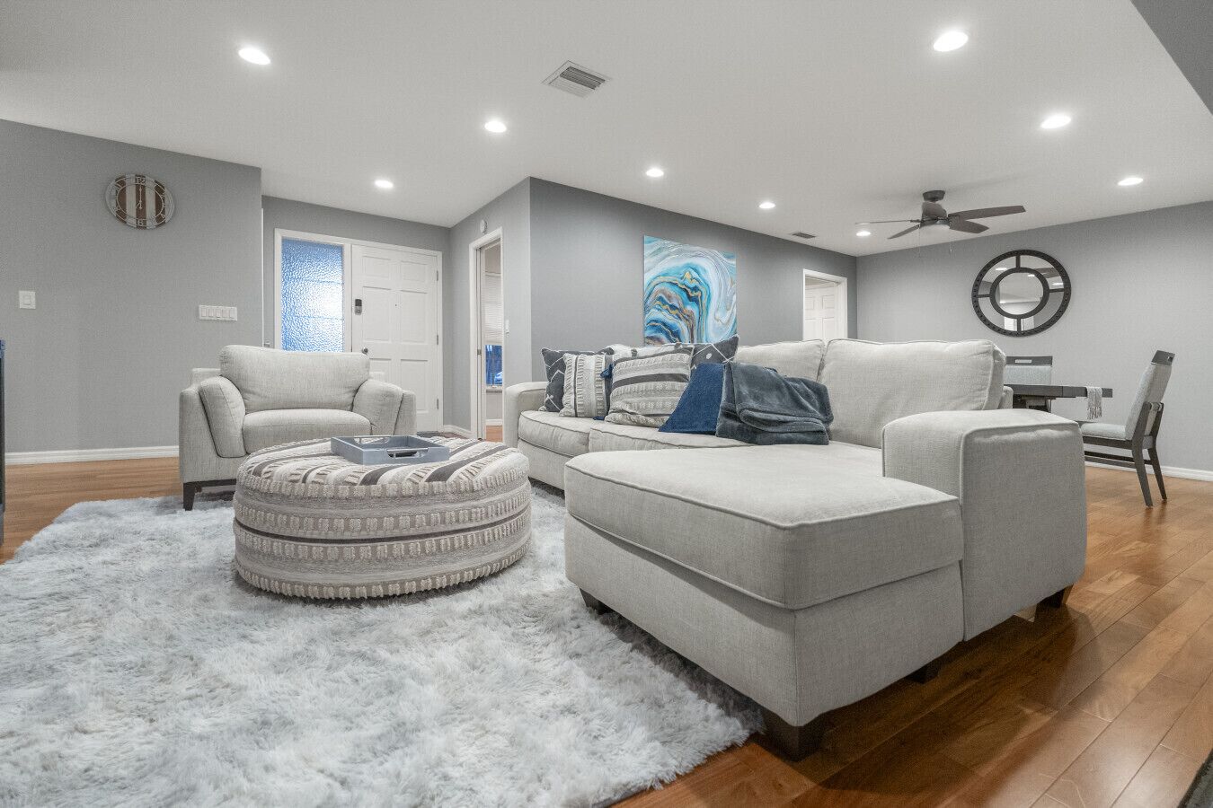 A modern living room with a grey sofa, ottoman, and armchair, a fluffy rug, and a ceiling fan.