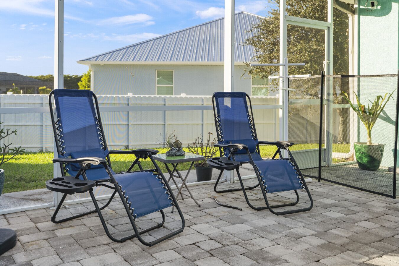 Eine Terrasse mit zwei blauen Klappstühlen und einem kleinen Tisch, einem weißen Zaun und einem Haus mit einem Metalldach im Hintergrund.
