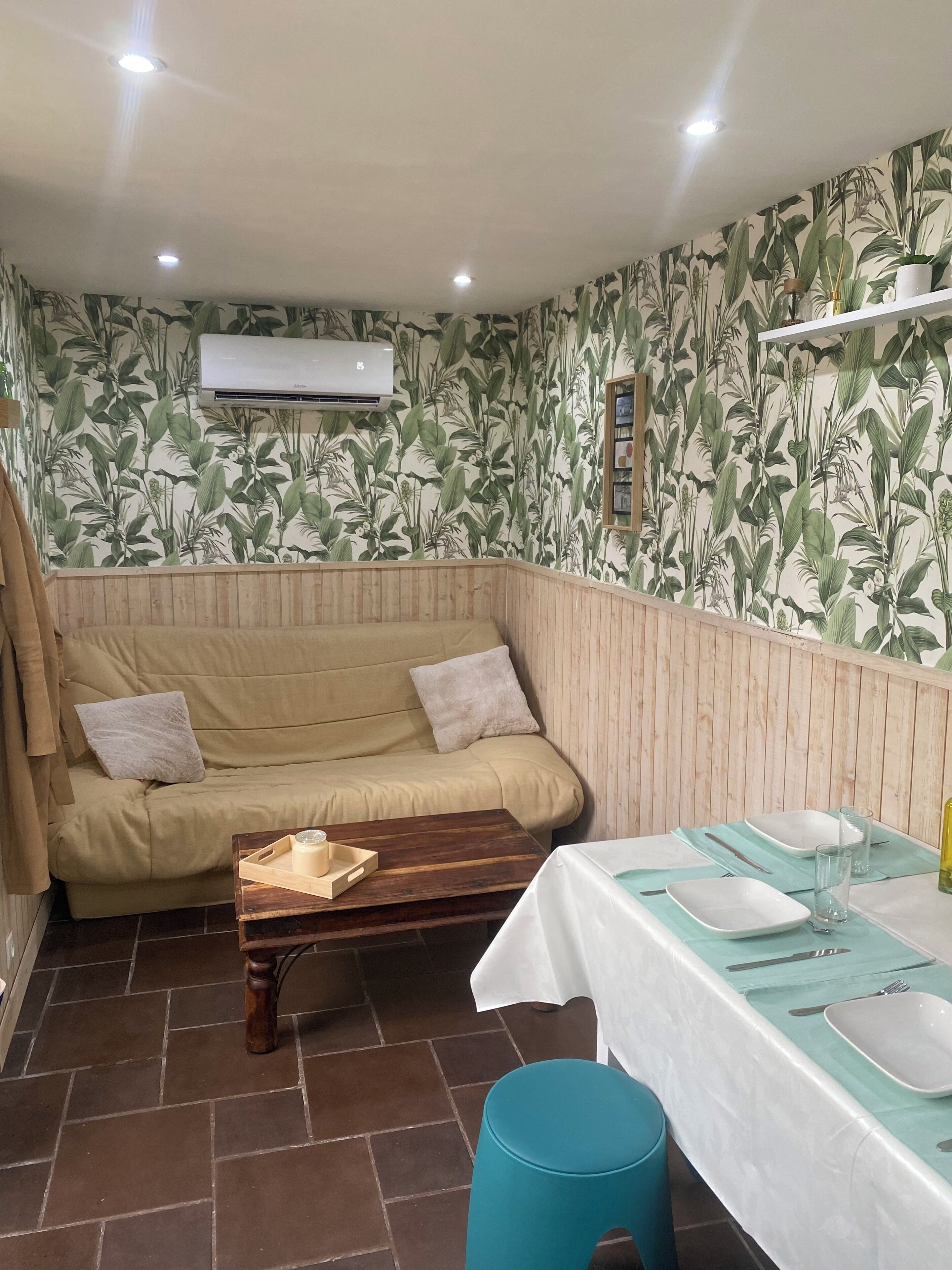 Una habitación con sofá color beige, una mesa de centro de madera, una mesa de comedor con mantel blanco y servilletas azules, y un aire acondicionado instalado en la pared.
