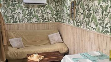 Una habitación con sofá color beige, una mesa de centro de madera, una mesa de comedor con mantel blanco y servilletas azules, y un aire acondicionado instalado en la pared.