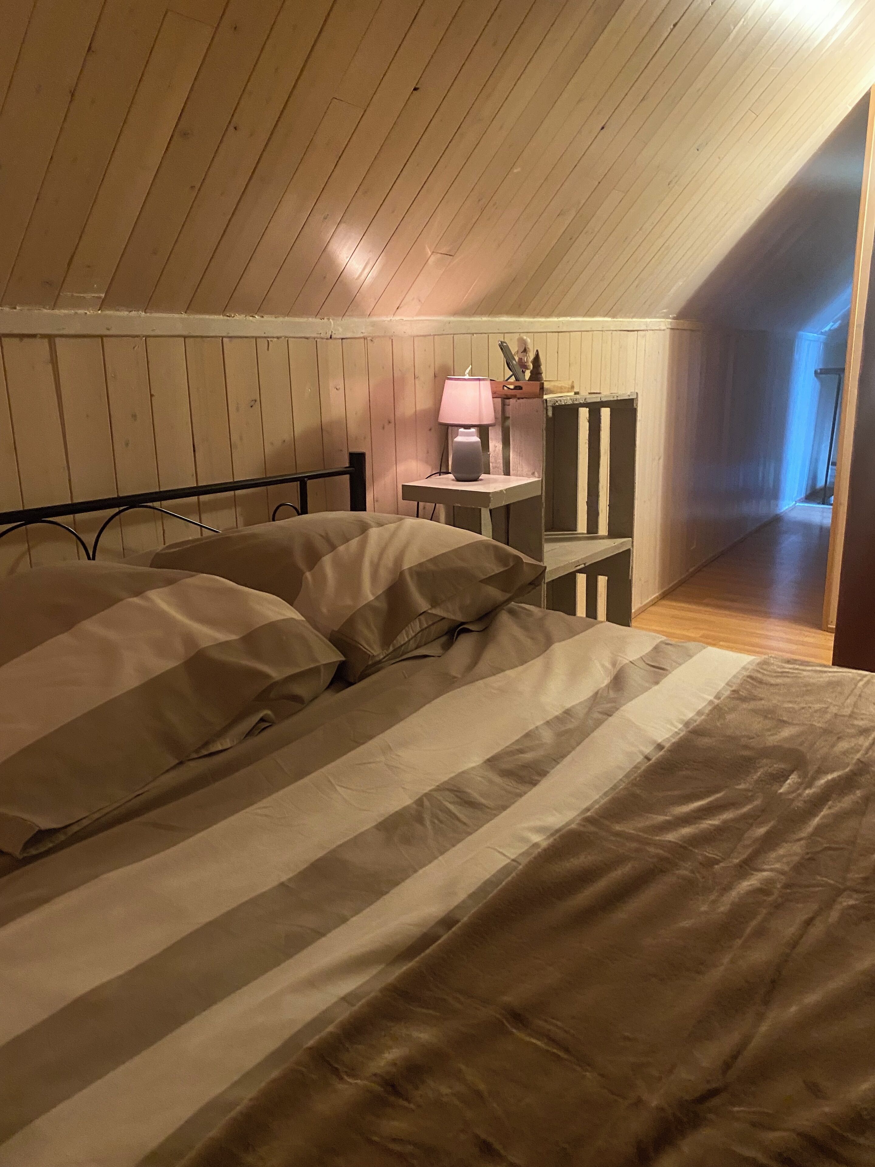 Un dormitorio con techo inclinado, una cama con ropa de cama a rayas, una mesita de noche con lámpara y un pequeño estante con adornos.