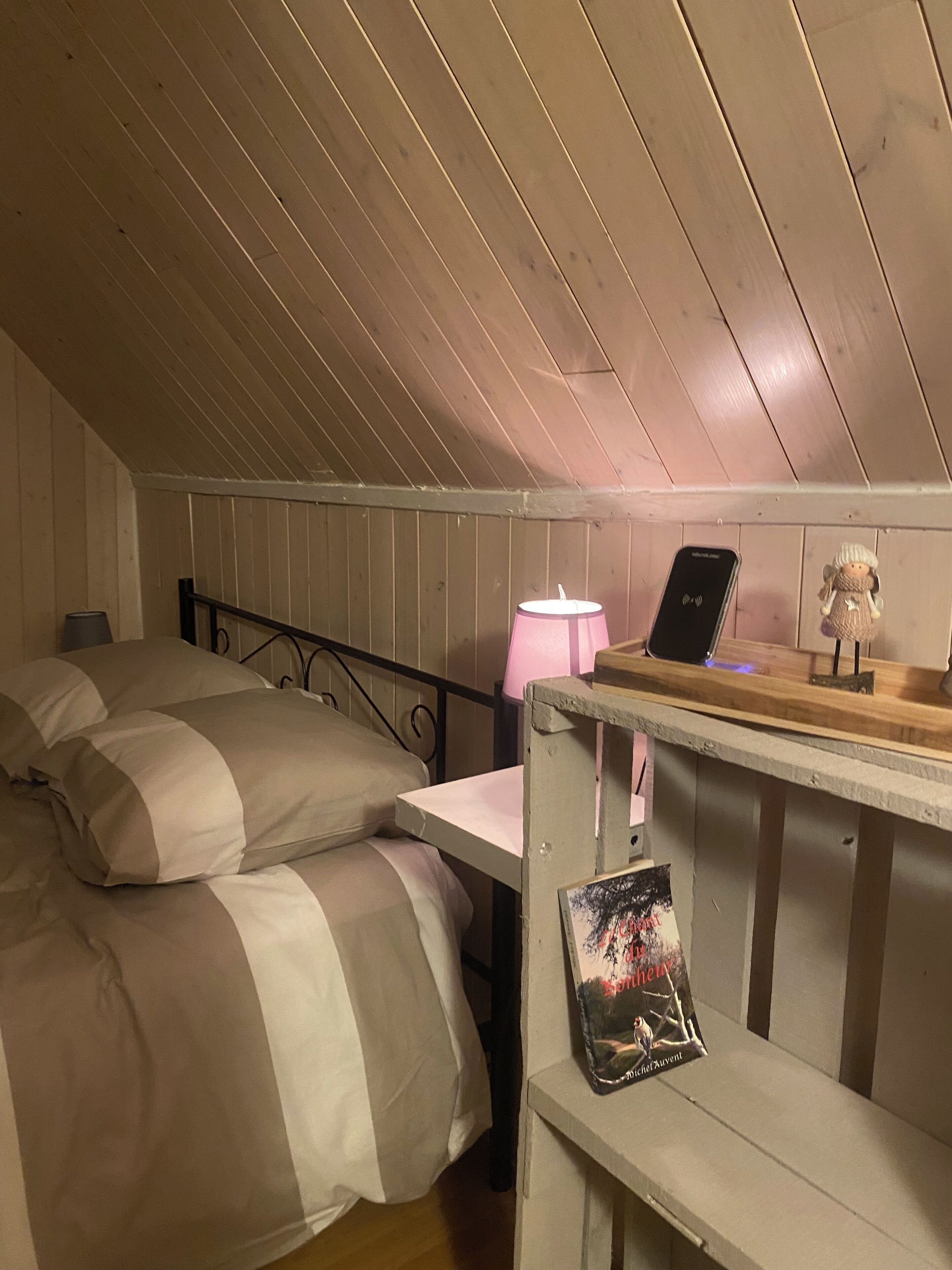 Ein kleines, gemütliches Schlafzimmer im Dachgeschoss mit schrägen Wänden, einem Einzelbett mit gestreifter Decke, einem Nachttisch mit Lampe und einem kleinen Regal mit einem Buch und einem Dekoartikel.