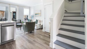 Uma cozinha moderna com máquina de lavar louça, um espaço de refeições com mesa e cadeiras, escadas e uma sala de estar com sofá e mesinha de centro.
