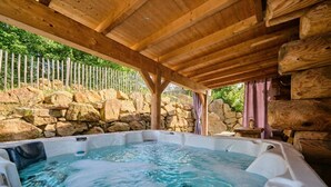 A hot tub under a wooden pergola with a stone wall and a fence in the background. - Log cabin chalet WoidSinn (Spiegelau)