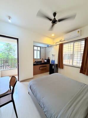 Une chambre avec un lit, un ventilateur de plafond, une fenêtre avec des rideaux, un coin cuisine et un balcon.