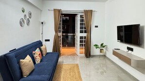 A modern living room with a blue sofa, a flat-screen TV, and a ceiling fan. - Osprey House: Modern AC 1BHK Stay by Bleisure (Pimpri-Chinchwad)