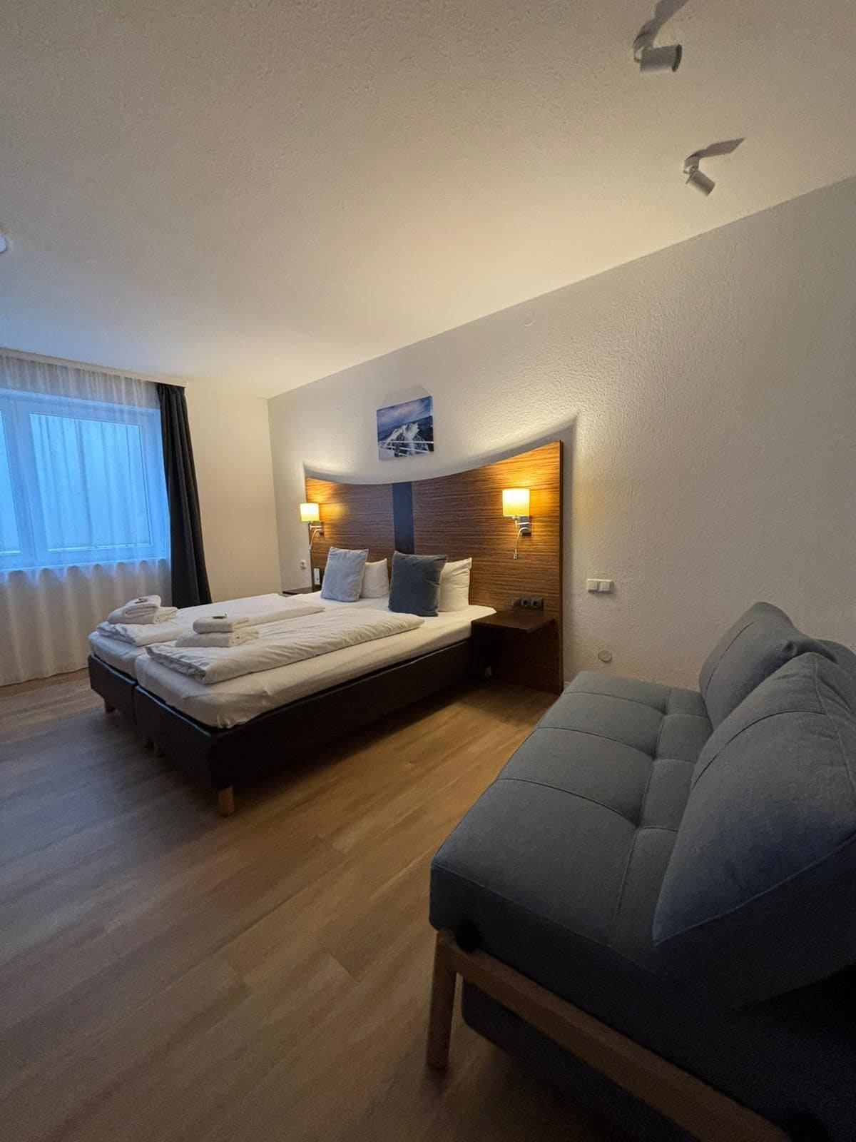 A hotel room with a bed, bedside lamps, a headboard with a picture, a sofa, and a window with curtains.