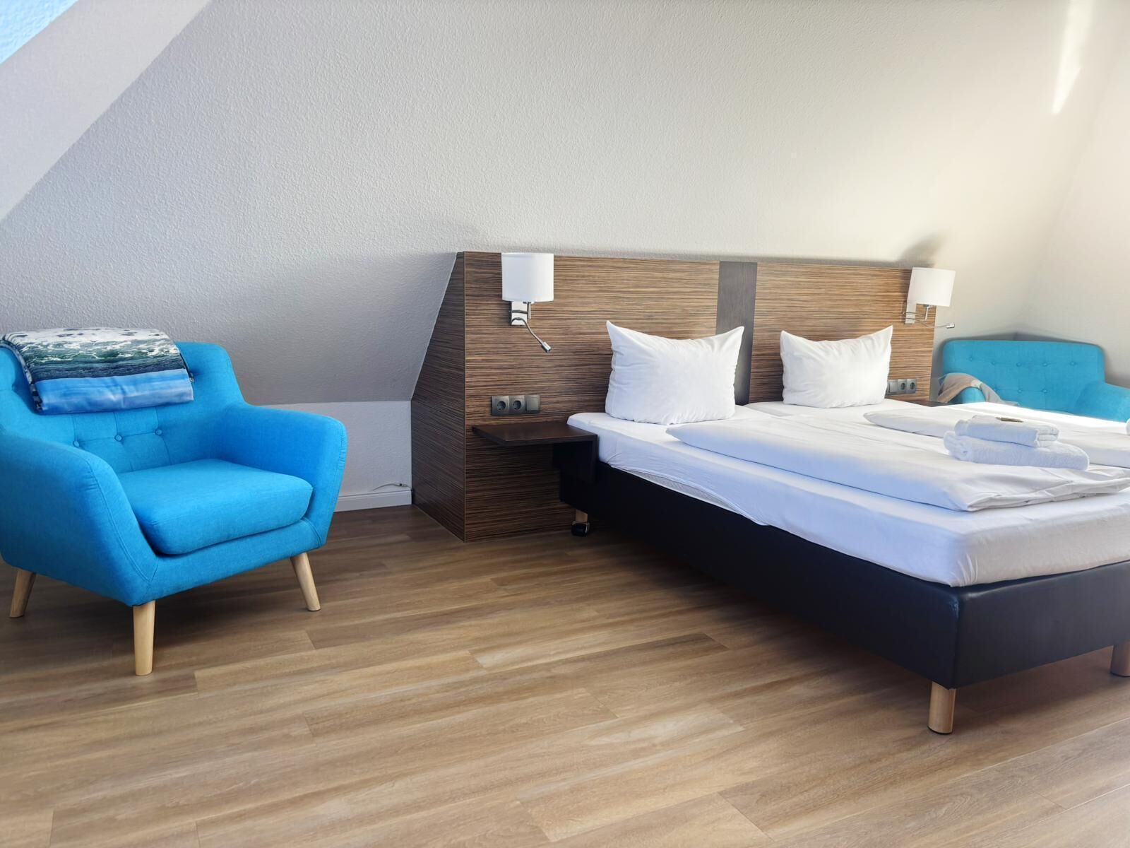Een hotelkamer met een bed, een blauwe fauteuil, een nachtkastje en een wandlamp.