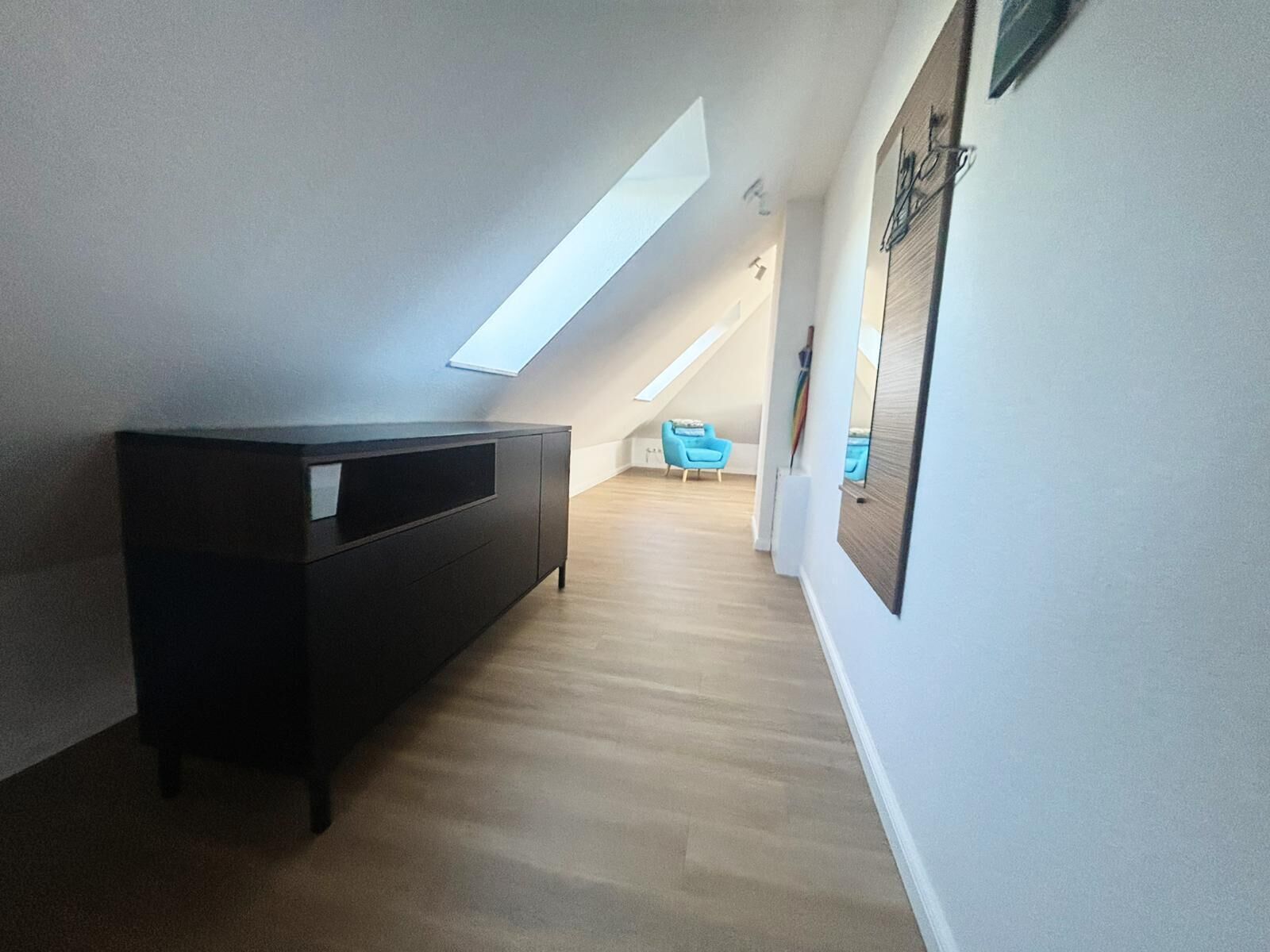 Un pasillo estrecho con un mueble oscuro, una silla azul y un techo inclinado con tragaluces.