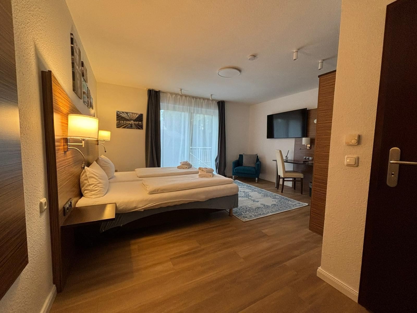Habitació d'hotel amb un llit, una taula de treball, una cadira, un televisor i una finestra amb cortines.