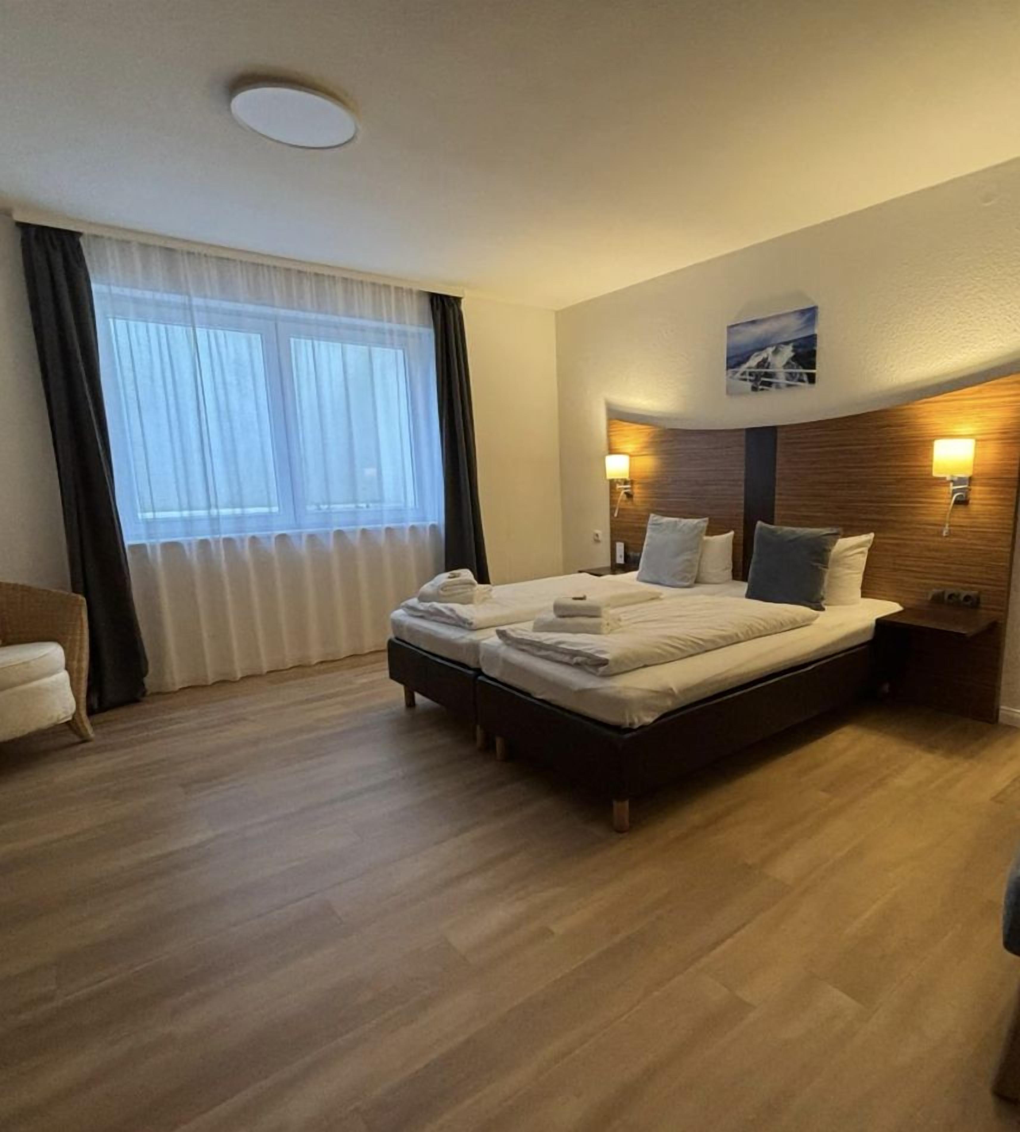 Ein Hotelzimmer mit einem Bett, Nachttischlampen, einem fensterbelaubten Fenster und Holzboden.