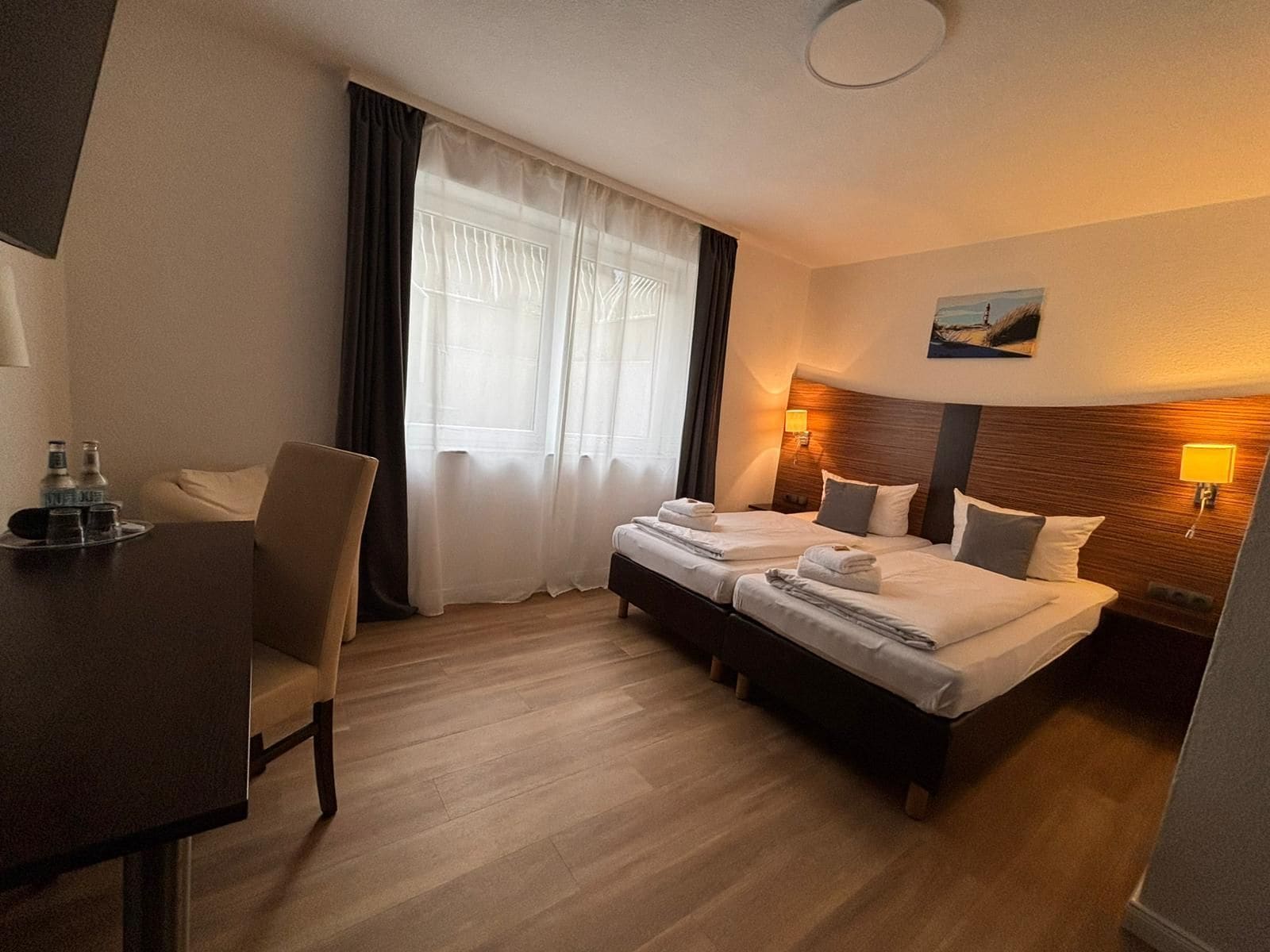 A hotel room with two beds, a desk, a chair, a window with curtains, and a painting on the wall.