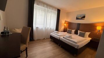 A hotel room with two beds, a desk, a chair, a window with curtains, and a painting on the wall.
