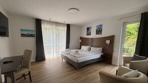 A hotel room with a bed, a desk, and a chair. There is a balcony with curtains and a view of greenery. - Turmhotel Scharbeutz (Scharbeutz)