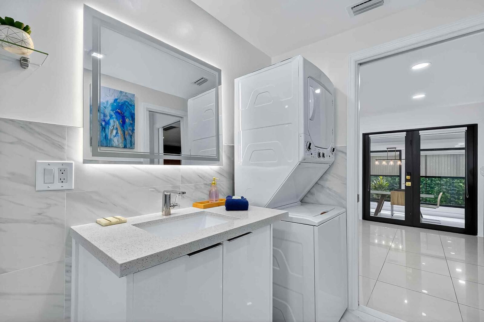 Une salle de bain moderne dotée d’un grand miroir, d’un lave-linge encastré et offrant une vue sur l’extérieur grâce à une porte vitrée.
