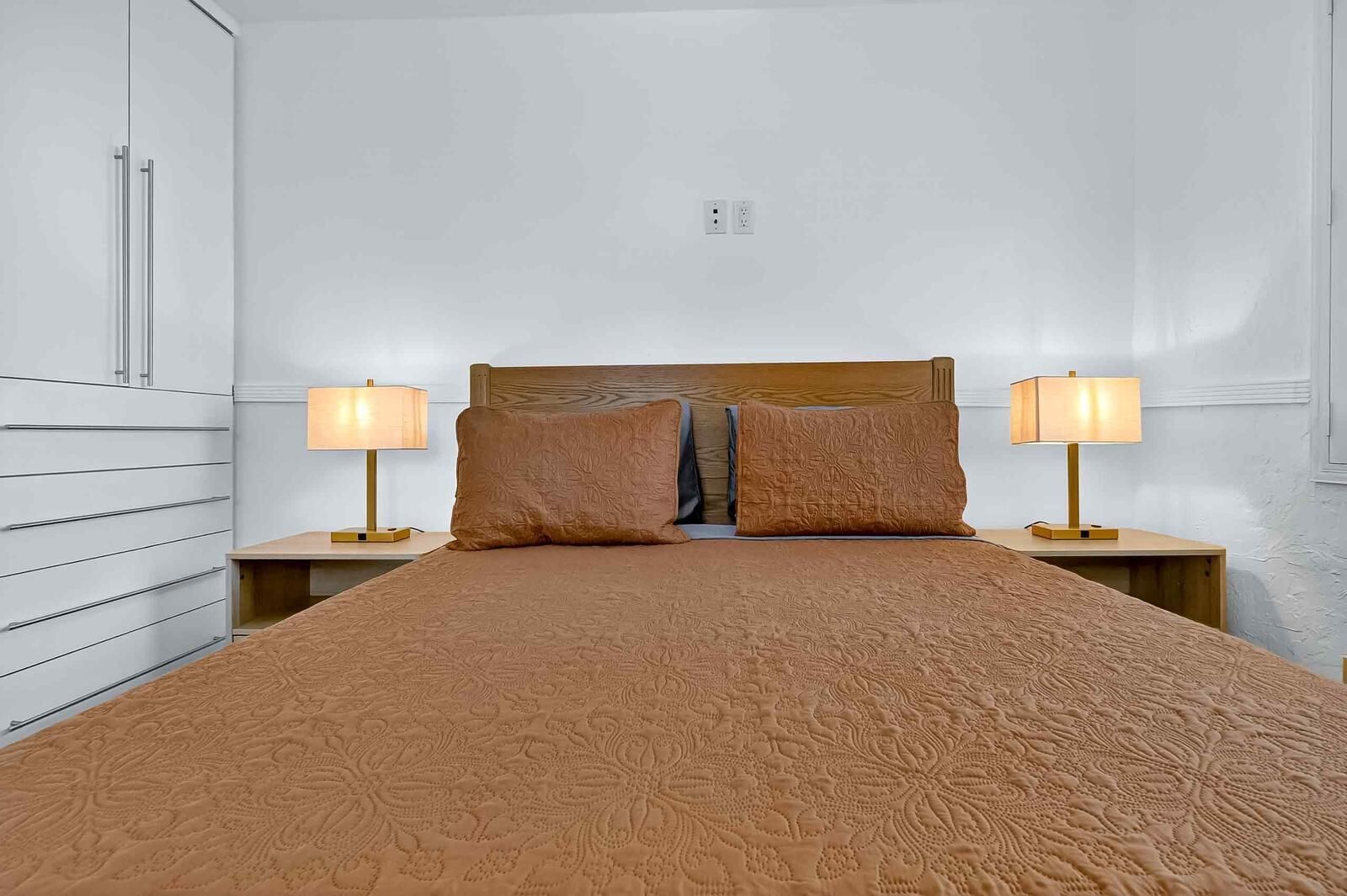 A neatly made bed with a patterned bedspread, flanked by two bedside lamps on wooden nightstands, against a plain wall with a power outlet.