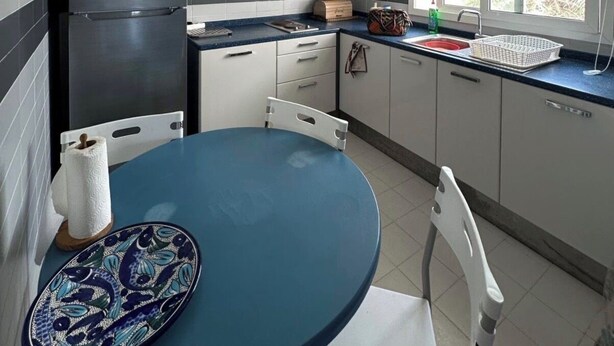 Eine kleine Küche mit grauen Schränken, einem schwarzen Kühlschrank, einem blauen Tisch und einem Fenster mit Jalousien.