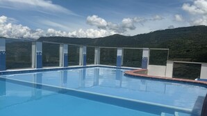 A clear blue pool with a tiled edge, surrounded by glass railings, set against a backdrop of green hills and a partly cloudy sky. - Suite 1103 (ideal for family travel) (San Gil)