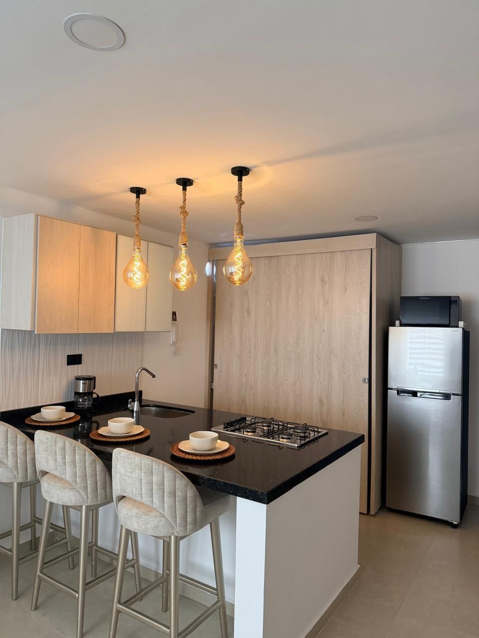 A modern kitchen with a central island, bar stools, a stainless steel refrigerator, and pendant lighting.