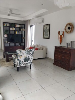 A living room with a sofa, a patterned armchair, a coffee table, a bookshelf, a TV, a fireplace, and a decorative wall piece. - Villa La Paisible de Palm Grove 2 (Grand Baie)