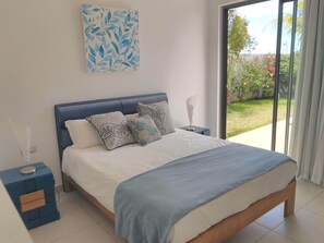 A bedroom with a bed, nightstands, a blue headboard, and a window with a view of greenery. - Villa La Paisible de Palm Grove 2 (Grand Baie)