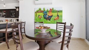 Zona pranzo con tavolo rotondo in vetro, quattro sedie e un quadro con galli sulla parete.
