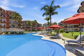 Een resort zwembad met ligbedden en parasols, omgeven door meerlaagsgebouwen met balkons.