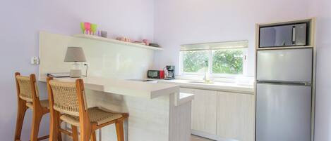 A kitchen with a white countertop, two wooden chairs, a refrigerator, and a window with a view of greenery.