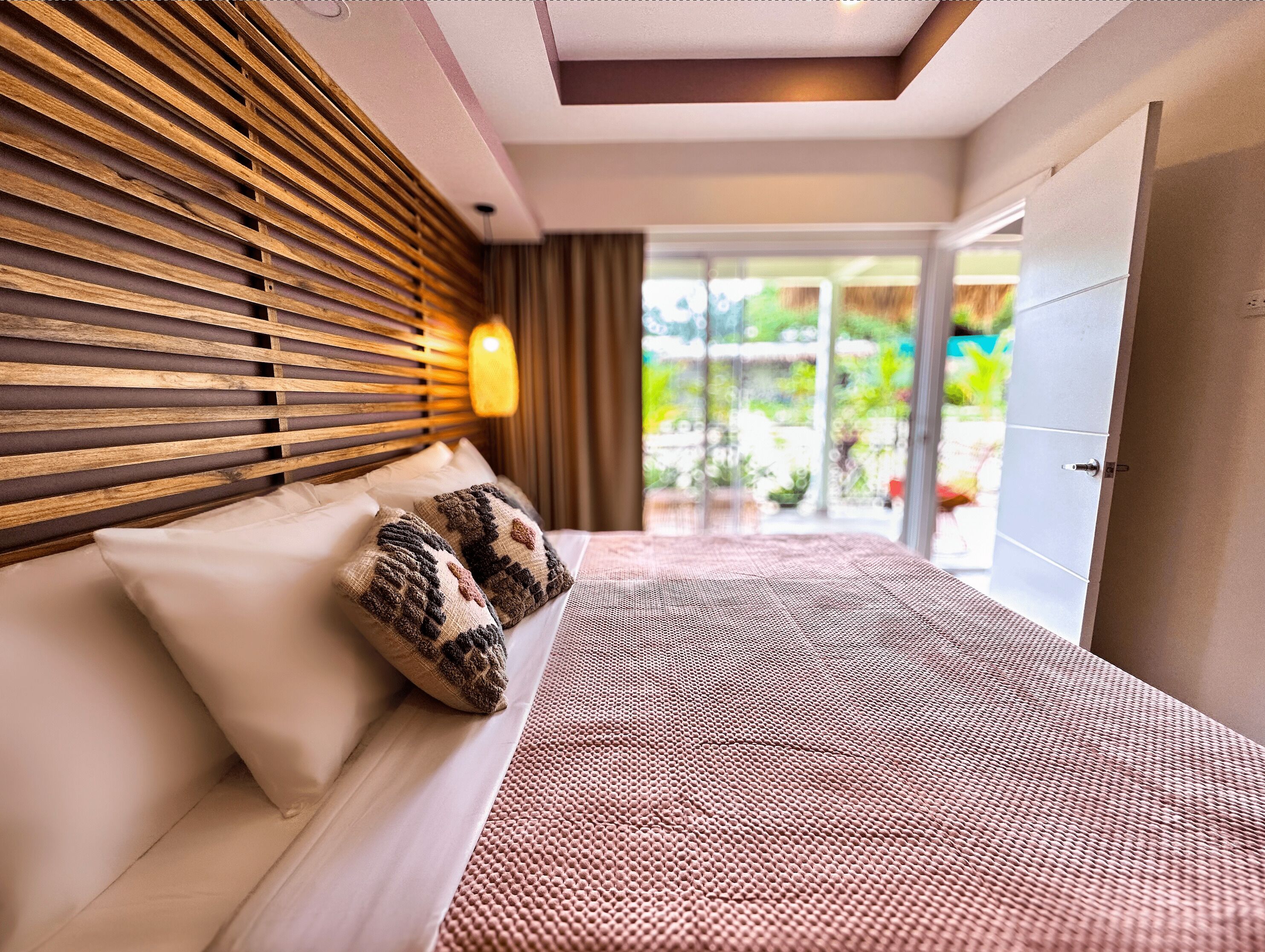 Un dormitorio moderno con un cabecero de madera con listones, una cama grande con ropa de cama estampada y vistas a un jardín a través de una puerta de vidrio.