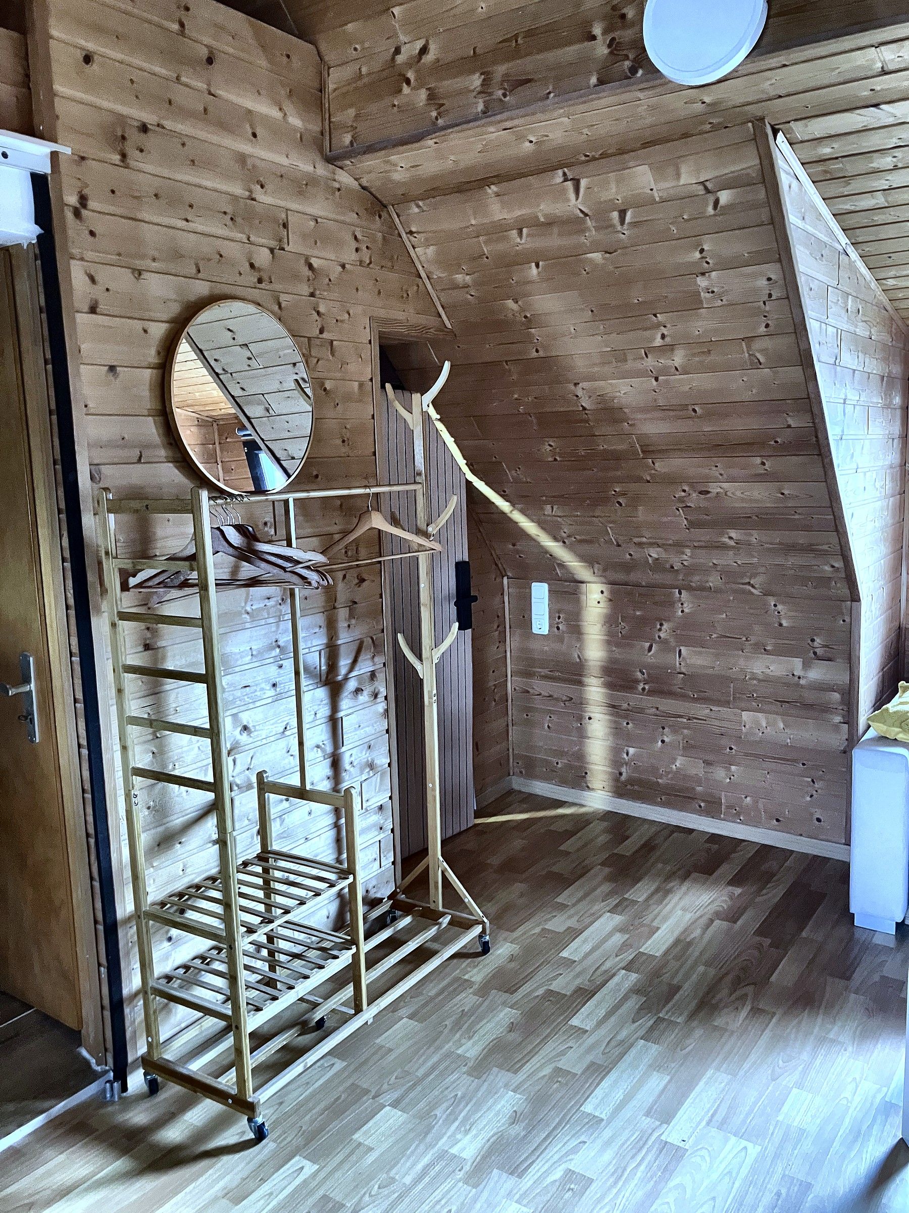 Une pièce aux murs en bois, un portant métallique avec des cintres, un miroir et une armoire blanche.