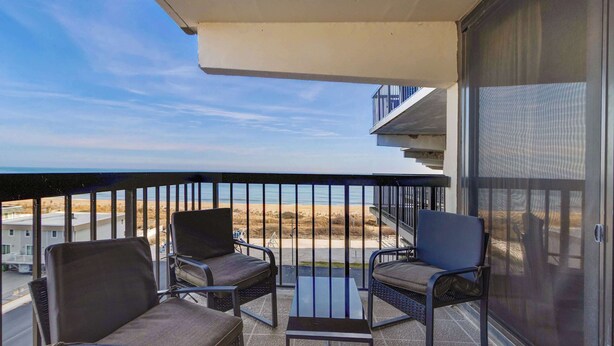 Ein Balkon mit zwei Stühlen und einem Tisch, mit Blick auf einen Strand und einen Parkplatz.