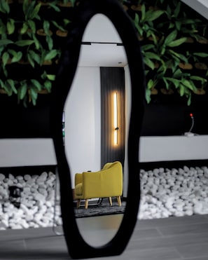 Un espejo refleja una habitación con un sillón amarillo, una cortina oscura y una pared cubierta de plantas.