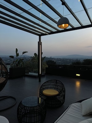 Terraço no terraço com vista para a paisagem urbana, apresentando uma pérgula, área de estar com cadeiras de vime e uma mesa de vidro.