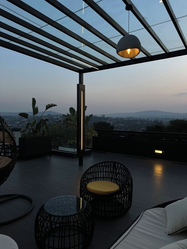 Una terraza en la azotea con vistas al paisaje urbano, que incluye una pérgola, un área de estar con sillas de mimbre y una mesa de vidrio.