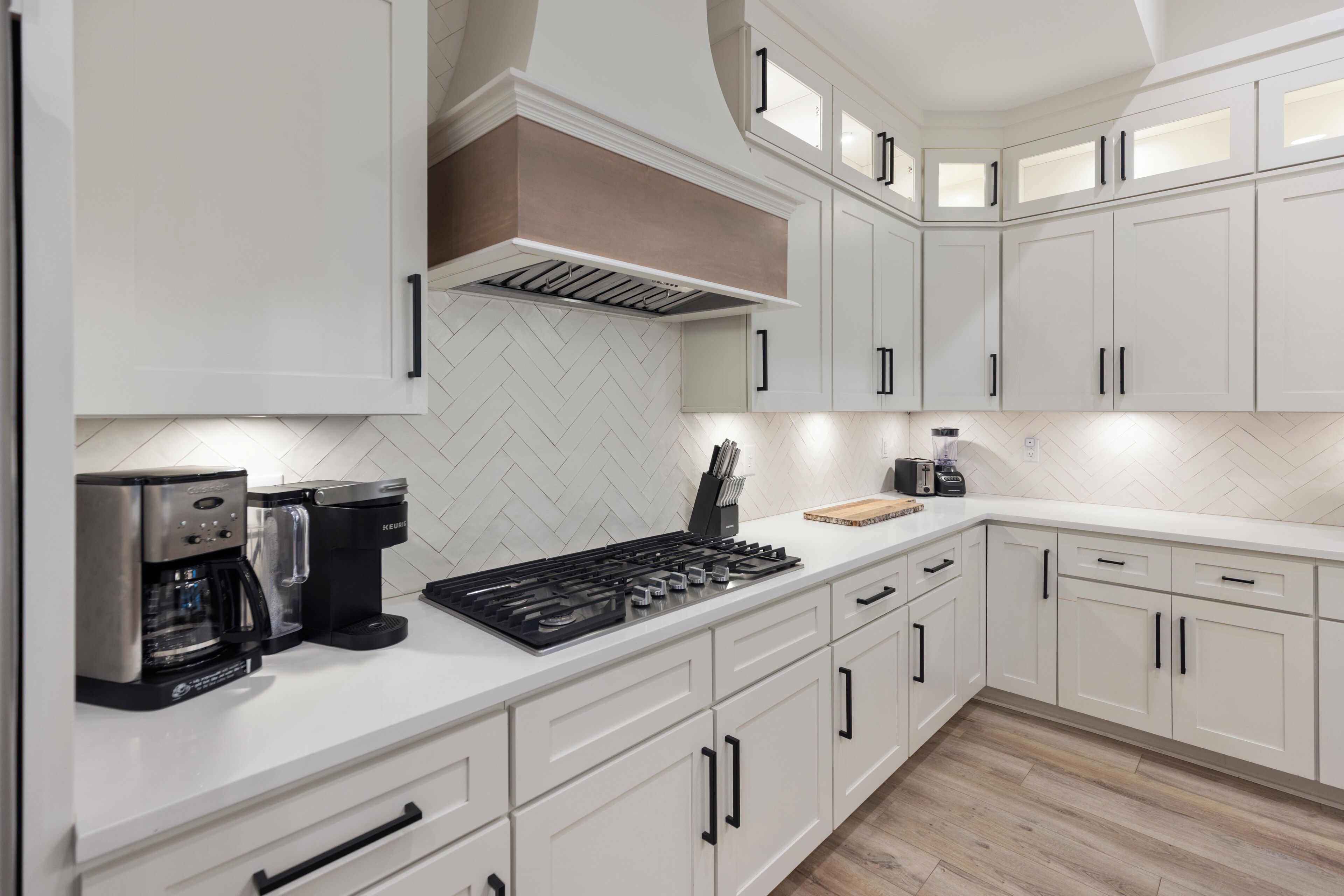 Une cuisine moderne avec des meubles blancs, un dosseret orné d’un motif en chevrons, une cuisinière encastrée et une machine à café.