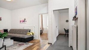 A modern living room with a grey sofa, a coffee table, and a rug. There is a hallway leading to another room with a bed and a chair.