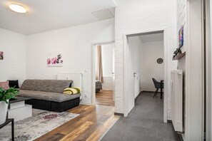 Una sala de estar moderna con un sofá gris, una mesa de centro y una alfombra. Se ve un pasillo que conduce a otra habitación con una cama y una silla.