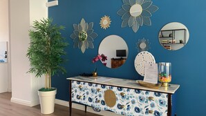 Ett rum med en blå vägg, ett vitt konsolbord med blommönster, en stor krukväxt och olika väggdekorationer, inklusive speglar och solstrålemotiv.