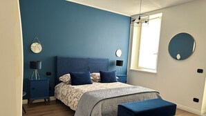 Ett sovrum med en blå accentvägg, en säng med blommigt sänglinne, ett sängbord med en lampa, en rund spegel och ett fönster som släpper in naturligt ljus.
