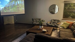 Olohuoneessa on suuri televisioruutu, kaksi nojatuolia, sohva, sohvapöytä ja seinällä peili.